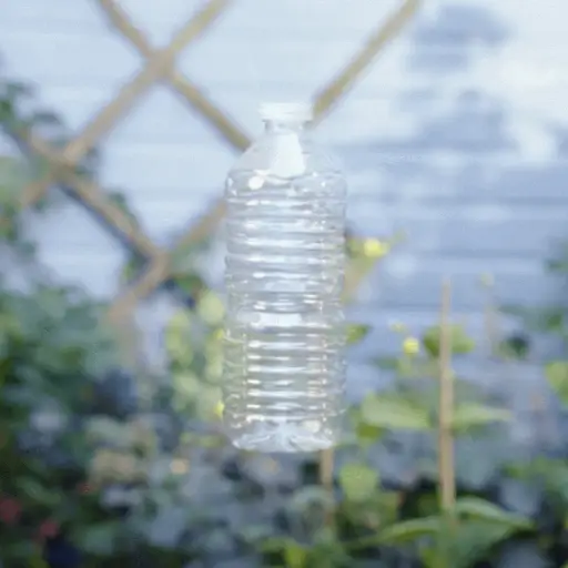 Clear plastic water bottle suspended in the air, with a slightly blurred garden background featuring green plants and a geometrically patterned fence.