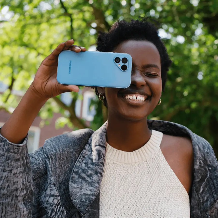 A smiling woman holding a Fairphone 4 in blue while posing outdoors.