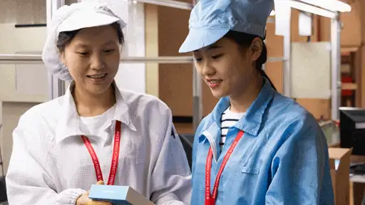 This image shows two factory workers wearing white and blue uniforms, holding a Fairphone box. The setting appears to be a factory or assembly line.